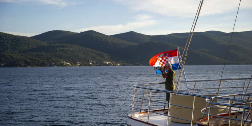 Reisender auf dem Deck eines Schiffes während einer Kreuzfahrt durch Kroatien – mit Blick auf die Küste und kroatischer Flagge am Heck.
